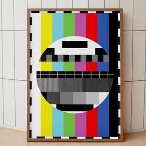 May include: A framed print with a classic television test card design. The artwork showcases vertical colour bars and a central circular element with black and grey patterns. The frame is a warm brown, adding a vintage feel.