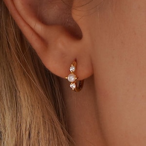 Puede incluir: Pendientes de aro dorados con una pequeña perla y dos cristales transparentes. Los pendientes se llevan en una oreja, destacando la joya. Los pendientes tienen un diseño delicado y elegante.