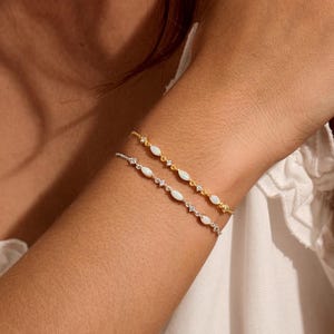 May include: Two delicate chain bracelets worn on the wrist, one in a gold tone and one in a silver tone. Each bracelet features a series of small, marquise-shaped white stones set at regular intervals along the thin, metallic link chain.
