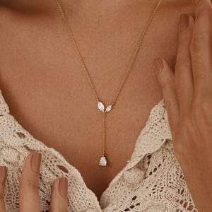 Könnte beinhalten: Eine goldfarbene Halskette mit einer filigranen Kette und drei weißen Edelsteinanhängern. Die Halskette hat ein Y-förmiges Design, mit zwei kleineren Edelsteinen oben und einem größeren tropfenförmigen Edelstein unten. Die Halskette wird von einer Person getragen.