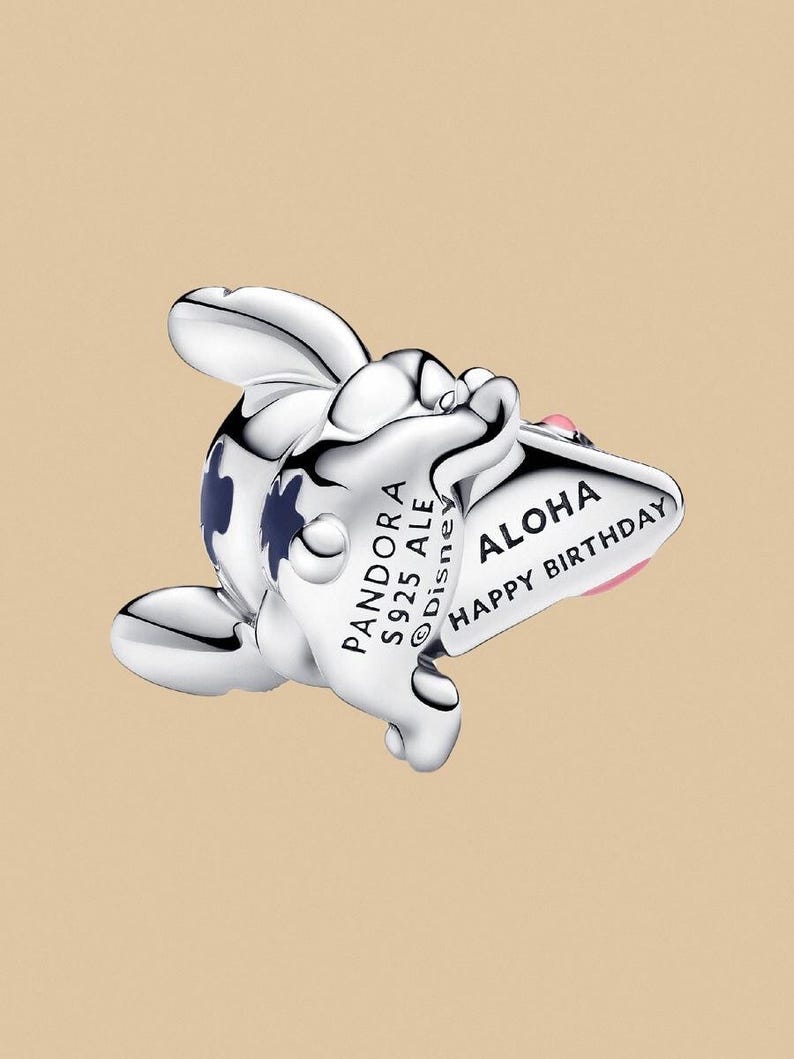 Puede incluir: Un charm de plata con un dise&ntilde;o de personaje de dibujos animados. El charm tiene un cuerpo blanco con detalles azules y un detalle rosa. Presenta el texto "ALOHA HAPPY BIRTHAY" en una pancarta y "PANDORA S925 ALE Disney" en el cuerpo.