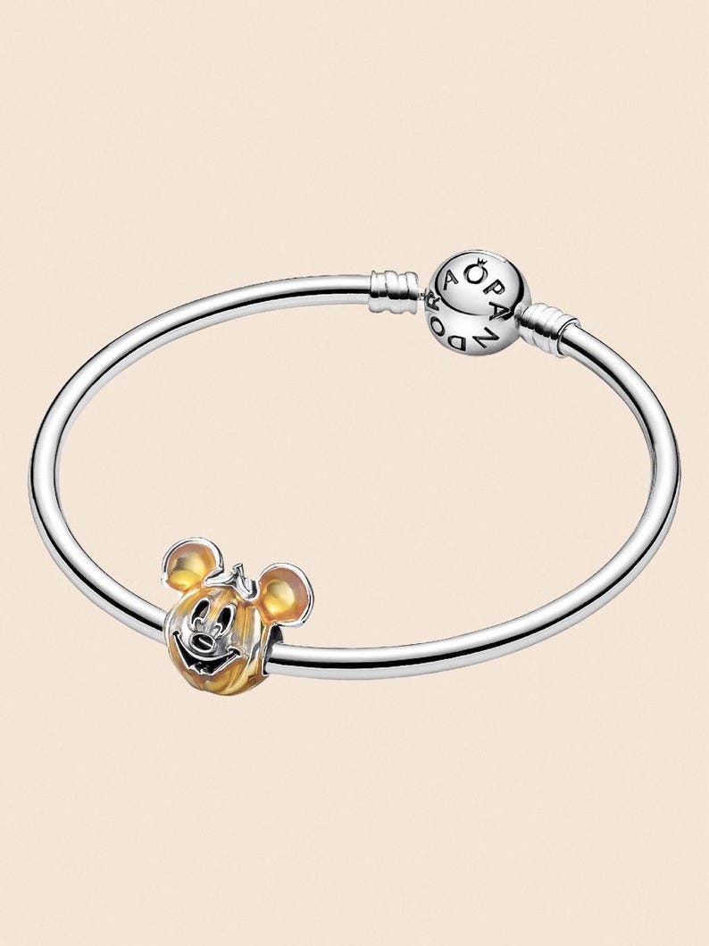 May include: A silver bracelet with a Mickey Mouse charm. The charm is a gold and silver Mickey Mouse head. The bracelet has a round clasp with the word "Pandora" on it.