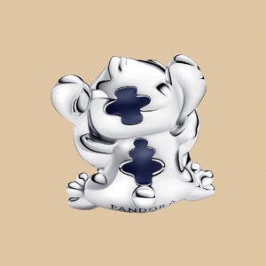 Puede incluir: Un charm plateado que representa un personaje de dibujos animados con grandes orejas y detalles en azul oscuro. El charm tiene un dise&ntilde;o en forma de cruz y el nombre de la marca es visible.