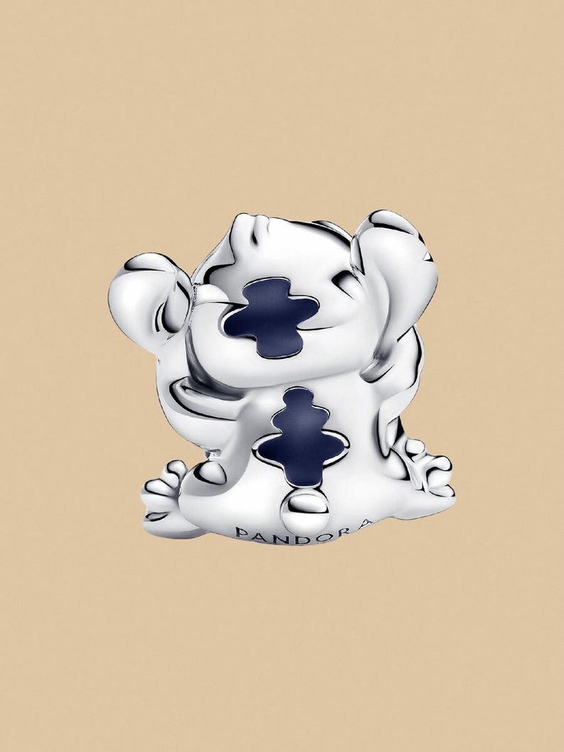 Puede incluir: Un charm plateado que representa un personaje de dibujos animados con grandes orejas y detalles en azul oscuro. El charm tiene un dise&ntilde;o en forma de cruz y el nombre de la marca es visible.