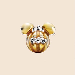 May include: A gold-colored charm shaped like a pumpkin with Mickey Mouse ears. The word "Boo" is displayed on the pumpkin. The charm has a shiny finish and is set against a light beige background. The charm is likely for a bracelet or necklace.