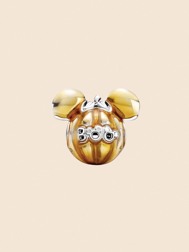 May include: A gold-colored charm shaped like a pumpkin with Mickey Mouse ears. The word "Boo" is displayed on the pumpkin. The charm has a shiny finish and is set against a light beige background. The charm is likely for a bracelet or necklace.