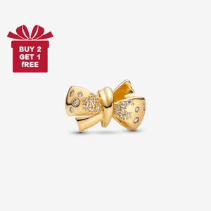Puede incluir: Un charm en forma de lazo dorado con piedras claras brillantes. El charm tiene un diseño detallado con un nudo en el centro y está sobre un fondo blanco. La imagen también incluye una caja de regalo roja con el texto "BUY 2 GET 1 FREE".