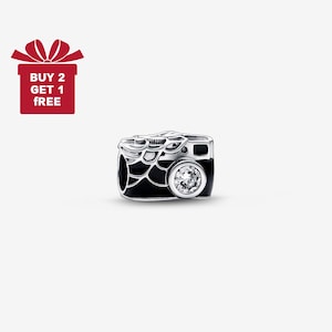 Peut inclure: Un charm argenté en forme d'appareil photo avec des détails en émail noir et une gemme ronde et transparente pour l'objectif. Le charm présente des détails complexes sur le dessus et les côtés. Un coffret cadeau rouge avec le texte "BUY 2 GET 1 FREE" est en haut à gauche.