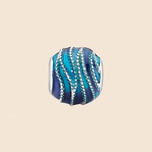 May include: A bead with a blue and teal swirl design, accented with white beaded lines. The bead has a silver-colored metal border. The design resembles an octopus tentacle. The bead is likely for jewelry making.