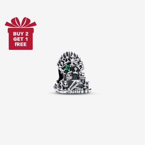 Puede incluir: Un charm de color plateado que representa un trono con una gema verde. El charm tiene detalles intrincados sobre un fondo blanco. Una caja de regalo roja con el texto "BUY 2 GET 1 FREE" está en la esquina superior izquierda.