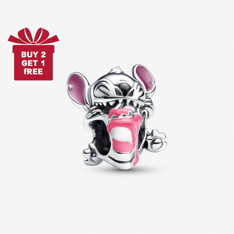 Puede incluir: Un charm plateado con el personaje Stitch de la pel&iacute;cula de Disney Lilo & Stitch. El charm tiene detalles rosas en las orejas y la lengua. La imagen tambi&eacute;n incluye una caja de regalo roja con el texto "BUY 2 GET 1 FREE".