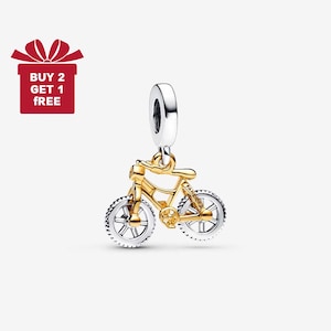 Puede incluir: Un charm de bicicleta dorado con ruedas plateadas y un lazo plateado para sujetarlo a una pulsera o collar. El charm es una réplica en miniatura de una bicicleta. Una caja de regalo roja con el texto "BUY 2 GET 1 FREE" está en la esquina superior izquierda.