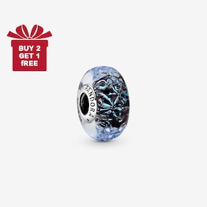 Puede incluir: Un charm de Pandora de cristal transparente con un diseño floral azul y negro. El charm tiene una banda plateada con el logo de Pandora. Una caja de regalo roja con el texto "BUY 2 GET 1 FREE" está en la esquina superior izquierda.
