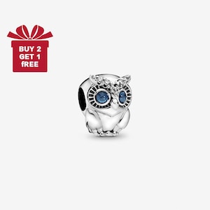 Puede incluir: Un charm de plata con forma de búho con ojos azules y una cabeza adornada con cristales. El charm tiene un diseño detallado y está sobre un fondo blanco. Una caja de regalo roja con el texto "BUY 2 GET 1 FREE" está en la esquina superior izquierda.