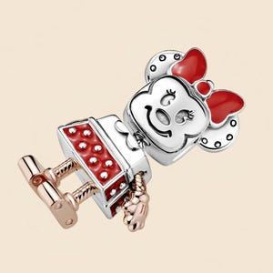 Pode incluir: Um berloque com um personagem de desenho animado com la&ccedil;o vermelho, vestido vermelho e rosto prateado. O berloque tem pernas e m&atilde;os em ouro rosa, inspirado em um personagem de desenho animado popular.