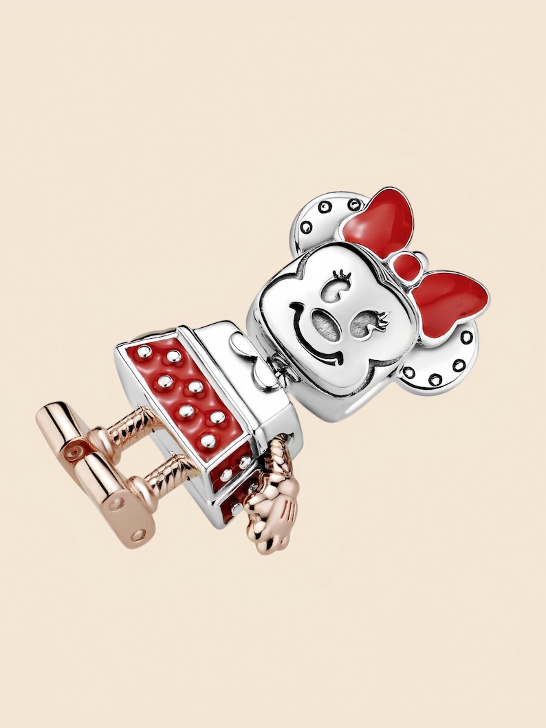 Pode incluir: Um berloque com um personagem de desenho animado com la&ccedil;o vermelho, vestido vermelho e rosto prateado. O berloque tem pernas e m&atilde;os em ouro rosa, inspirado em um personagem de desenho animado popular.