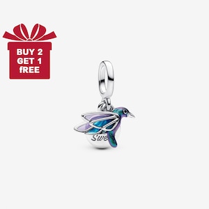 Puede incluir: Un charm de plata con un diseño de colibrí. El pájaro tiene alas y cuerpo de esmalte morado, azul y verde. El charm está suspendido de un lazo plateado. La palabra "Sweet" está escrita en la parte inferior del charm.