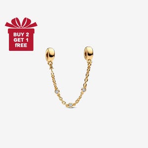 Può includere: Un charm di sicurezza a catena color oro con tre pietre preziose rotonde e trasparenti. Il charm ha due estremità arrotondate e una catena che le collega. Un cofanetto rosso con la scritta "BUY 2 GET 1 FREE" si trova nell'angolo in alto a sinistra.