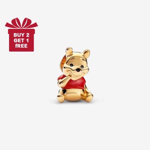 May include: A gold-coloured Winnie the Pooh charm wearing a red shirt. The charm is a miniature figurine of the beloved character. The image also includes a red gift box with the text "BUY 2 GET 1 FREE".