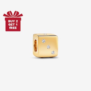 May include: A gold-colored charm in the shape of a die, featuring three clear, sparkling gemstones. The charm has a smooth, polished surface and a hole for threading onto a bracelet or necklace. A red gift box with the text "BUY 2 GET 1 FREE" is in the image.