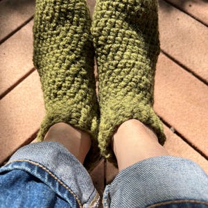 May include: A pair of green crocheted slippers with a textured pattern. The slippers are worn by a person wearing blue jeans.