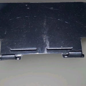 May include: A black plastic handheld gaming controller grip featuring ergonomic side handles with a textured surface, a central rectangular mounting area, and two small bottom support clips designed to hold a portable gaming device securely.