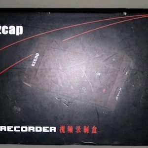 May include: A black rectangular product box for an ezcap AV RECORDER. The box features the white text ezcap in the top left corner, two thin red diagonal lines, and the text AV RECORDER with Chinese characters printed at the bottom edge.