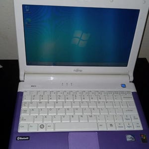 Puede incluir: Un portátil Fujitsu M2010 blanco y morado con teclado QWERTY, panel táctil y una pantalla que muestra un fondo de escritorio Windows azul. El dispositivo cuenta con una cámara web en el marco superior y marcas de Bluetooth e Intel Atom.