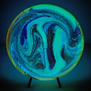 Op de afbeelding: Een rond, abstract kunstwerk met een wervelend patroon van blauw, groen en bruin. Het kunstwerk is gemonteerd op een kleine, donkere metalen standaard. De kleuren gloeien met een etherische kwaliteit, waardoor een uniek decoratief item ontstaat. Diameter ca. 20 cm.