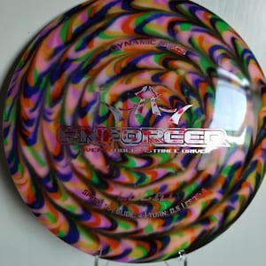 May include: A disc golf disc with a vibrant, swirled design in shades of pink, orange, green, blue, and purple. The disc features the text "ENFORCER" and "DYNAMIC DISCS". It is a versatile distance driver.