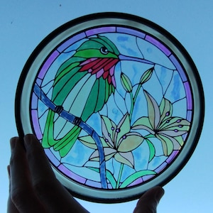 May include: A stained glass window panel featuring a hummingbird with a long beak, perched on a branch with two white lilies. The hummingbird is green, red, and blue, while the lilies are white and yellow. The background is a light blue.