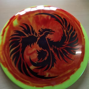 Könnte beinhalten: Eine Disc-Golf-Scheibe mit einem lebendigen Design. In der Mitte befindet sich eine schwarze Phönix-Silhouette vor einem wirbelnden Hintergrund aus Rot, Orange und Gelb. Der äußere Rand ist hellgrün.