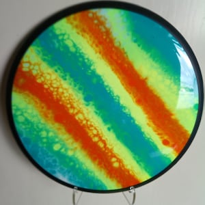 Könnte beinhalten: Runde Wandkunst mit schwarzem Rahmen. Das Kunstwerk zeigt ein lebendiges abstraktes Design mit diagonalen Streifen in Türkis, Gelb und Orange. Die Farben verschmelzen zu einem einzigartigen, auffälligen Stück.