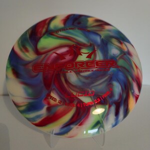 May include: A multi-coloured disc golf disc with a swirl pattern in shades of red, blue, green, and white. The disc has the words "Dynamic Discs" and "Enforcer" printed on it in red. The disc has the following statistics printed on it: "Speed: 12 | Glide: 4 | Turn: 0.5 | Fade: 1"