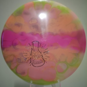 Op de afbeelding: Een kleurrijke discgolfschijf met een levendige mix van roze, oranje en groene tinten. De schijf heeft een zwarte afbeelding van een beker met de tekst "Lab Second". De schijf wordt op een transparante standaard gepresenteerd.