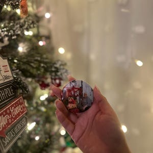May include: A round Christmas ornament with a photo of two people in front of a Christmas tree. The ornament is red and white with a gold trim.