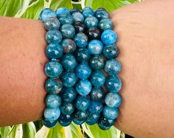 May include: A collection of blue apatite gemstone bracelets. The bracelets are made of round, polished beads in shades of teal and turquoise. The bracelets are worn on a wrist, with green and white foliage in the background.