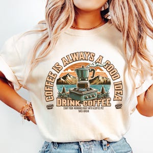 May include: Cream-colored t-shirt with a circular graphic that reads "Coffee is Always a Good Idea." The graphic features a coffee pot, mountains, and the words "Drink Coffee." The shirt has short sleeves and a relaxed fit.