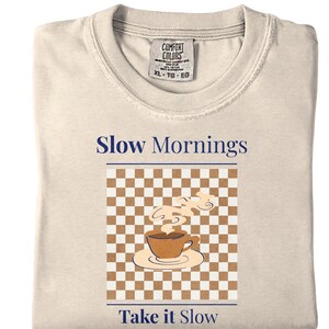 May include: Cream-colored t-shirt with the words "Slow Mornings" and "Take it Slow" in navy blue. The shirt features a graphic of a coffee cup on a saucer with steam rising, set against a brown and white checkered background. The tag reads XL-TG-EG.