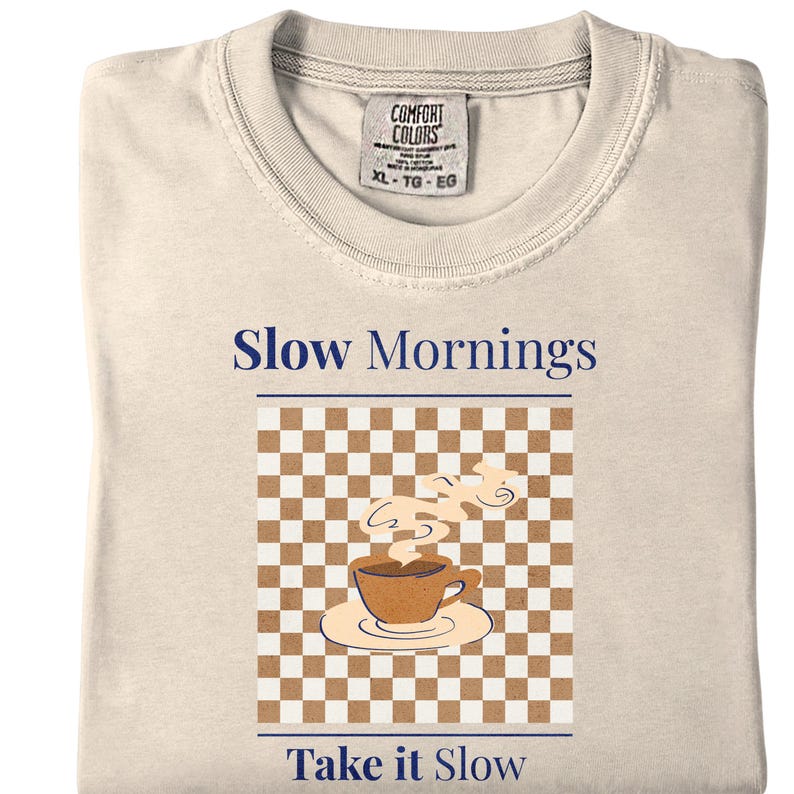 May include: Cream-colored t-shirt with the words "Slow Mornings" and "Take it Slow" in navy blue. The shirt features a graphic of a coffee cup on a saucer with steam rising, set against a brown and white checkered background. The tag reads XL-TG-EG.