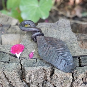 Pode incluir: Um pingente de metal cinza-escuro forjado à mão em forma de folha. O topo do pingente apresenta um laço de metal torcido para pendurar. A superfície da folha exibe detalhes de veios lineares gravados em todo o seu corpo metálico.