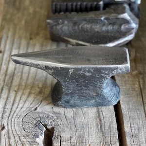 May include: A small, dark grey metal anvil with a smooth, polished top surface and a pointed horn. The body of the anvil features a textured, forged finish with visible hammer marks, showcasing a sturdy, compact design for metalworking tasks.
