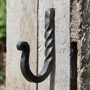 May include: A black metal wall hook featuring a twisted vertical shaft and a curved, scrolled end. The hook is mounted onto a vertical wooden surface, displaying a dark, textured finish with visible hammer marks along the metal structure.