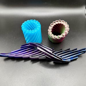 Op de afbeelding: Drie abstracte decoratieve objecten. Een lichtblauwe, spiraalvormige cilinder, een veelkleurige cilinder met een geschulpte rand en een reeks metalen, iriserende, vleugelachtige vormen in tinten paars en blauw. De objecten worden op een donkere ondergrond getoond.