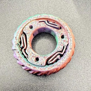 Puede incluir: Objeto circular multicolor con superficie texturizada y agujero central. El objeto presenta un diseño en capas con una mezcla de colores, incluyendo rojo, verde y morado. El borde exterior tiene un patrón estriado y varios agujeros más pequeños.