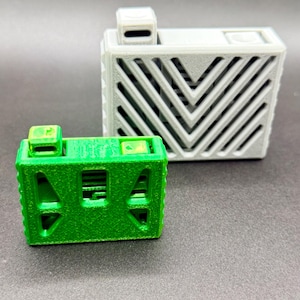 Puede incluir: Dos cajas geométricas impresas en 3D. Una es de color verde brillante con forma rectangular y un pequeño cuadrado en la parte superior. La otra es gris, más grande, y tiene forma rectangular con un patrón de chevrón y un pequeño cuadrado en la parte superior.
