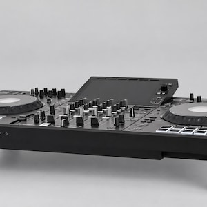 Pode incluir: Um controlador de DJ Pioneer preto com dois jog wheels circulares, uma secção de mistura central com vários botões e faders, um ecrã superior e pads de desempenho iluminados, tudo apoiado sobre quatro pés cilíndricos pretos.