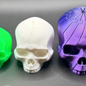 Puede incluir: Tres figuras decorativas de calaveras en varios tamaños y colores. La más pequeña es verde brillante, la del medio es blanca y la más grande es morada y azul. Cada calavera tiene rasgos faciales detallados y un diseño único en la parte superior.