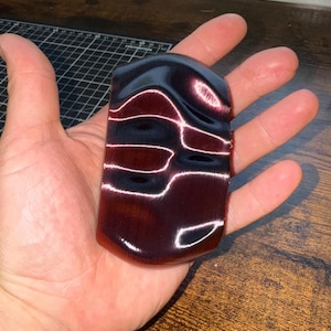 May include: A rectangular, dark burgundy object with a glossy finish. The object has a curved top and is accented with white, wavy lines. The item is held in an open hand, showcasing its shape and colour.