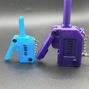 Könnte beinhalten: Zwei 3D-gedruckte Schlüsselanhänger in Form von Miniatur-Walkie-Talkies. Einer ist hellblau, der andere ist lila. Beide tragen den Text "FD M57". Jeder Schlüsselanhänger hat eine kleine Metallkette.